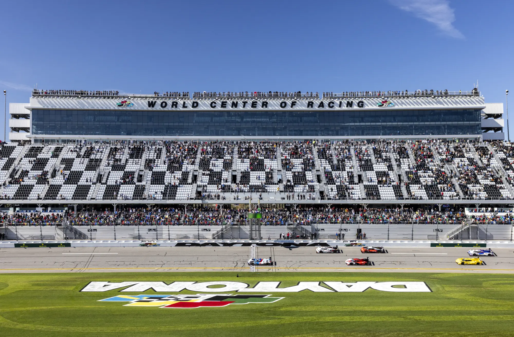 Szerokie ujęcie toru Daytona International Speedway z zatłoczoną trybuną pod czystym, błękitnym niebem i samochodami ścigającymi się na torze.