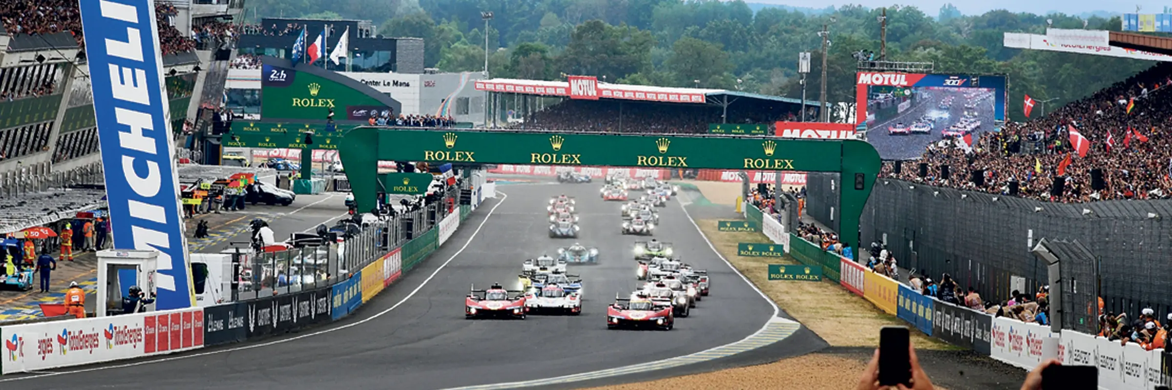 Wyścig samochodów obok kultowego łuku Rolex na starcie 24-godzinnego wyścigu Le Mans, z entuzjastycznym tłumem i widocznym wsparciem zespołu.